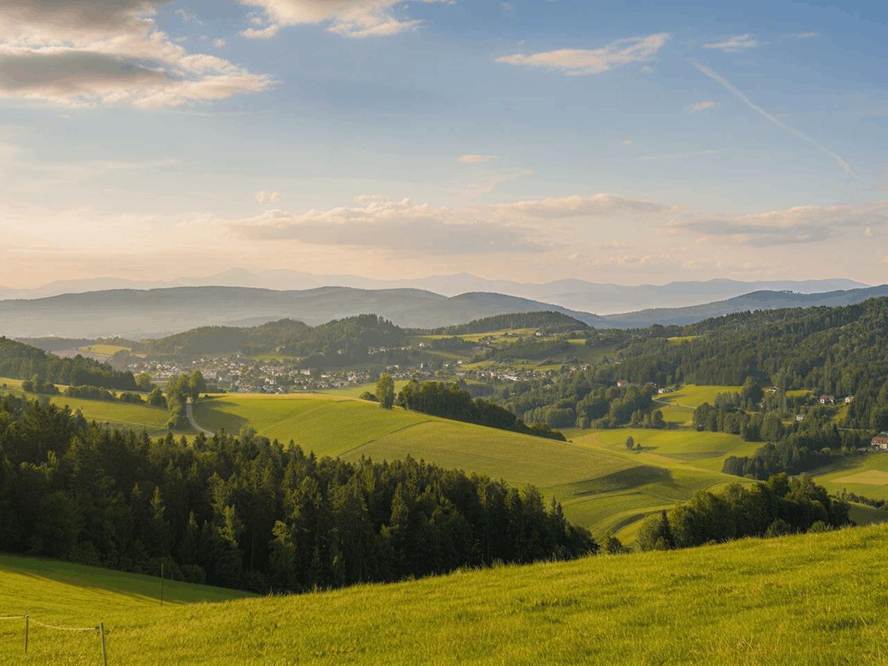 Weite Hügellandschaft mit grünen Wiesen, Wäldern und einem Dorf im Tal, im Hintergrund sanfte Berge unter blauem Himmel mit Wolken.